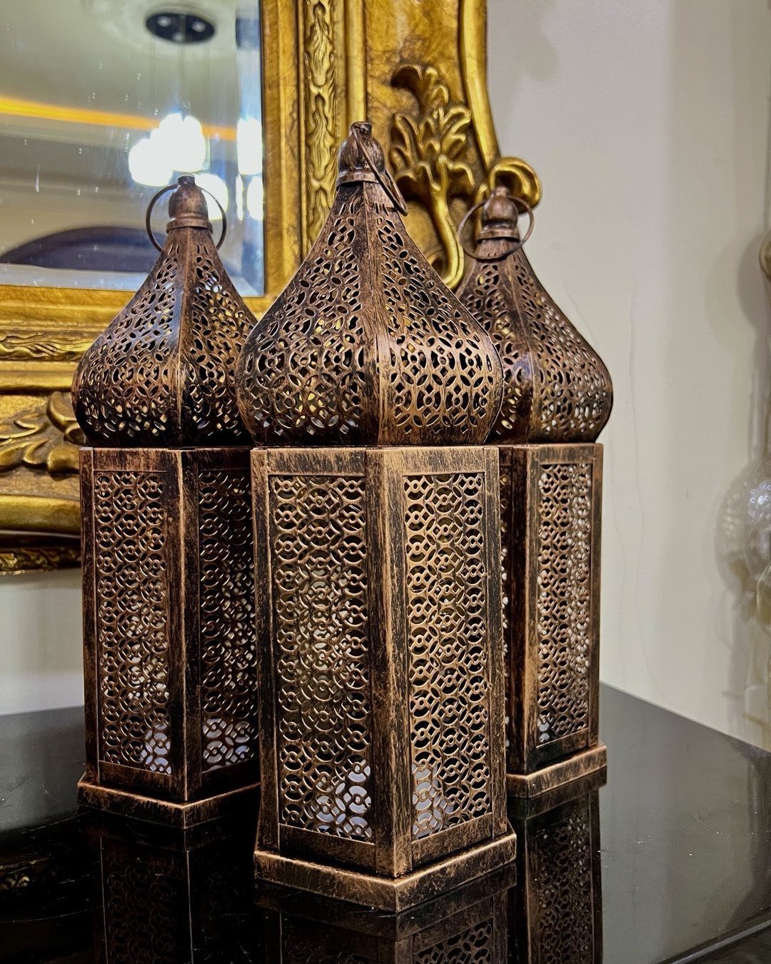Three bronze metal lanterns with intricate geometric patterns sitting on a reflective dark surface.