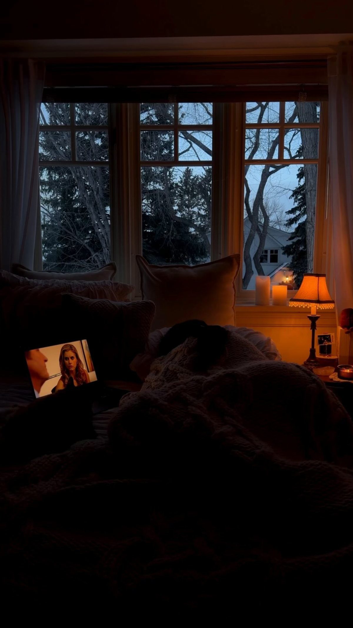A cozy bed with textured pillows and a shaggy faux-fur blanket near a window at dusk.