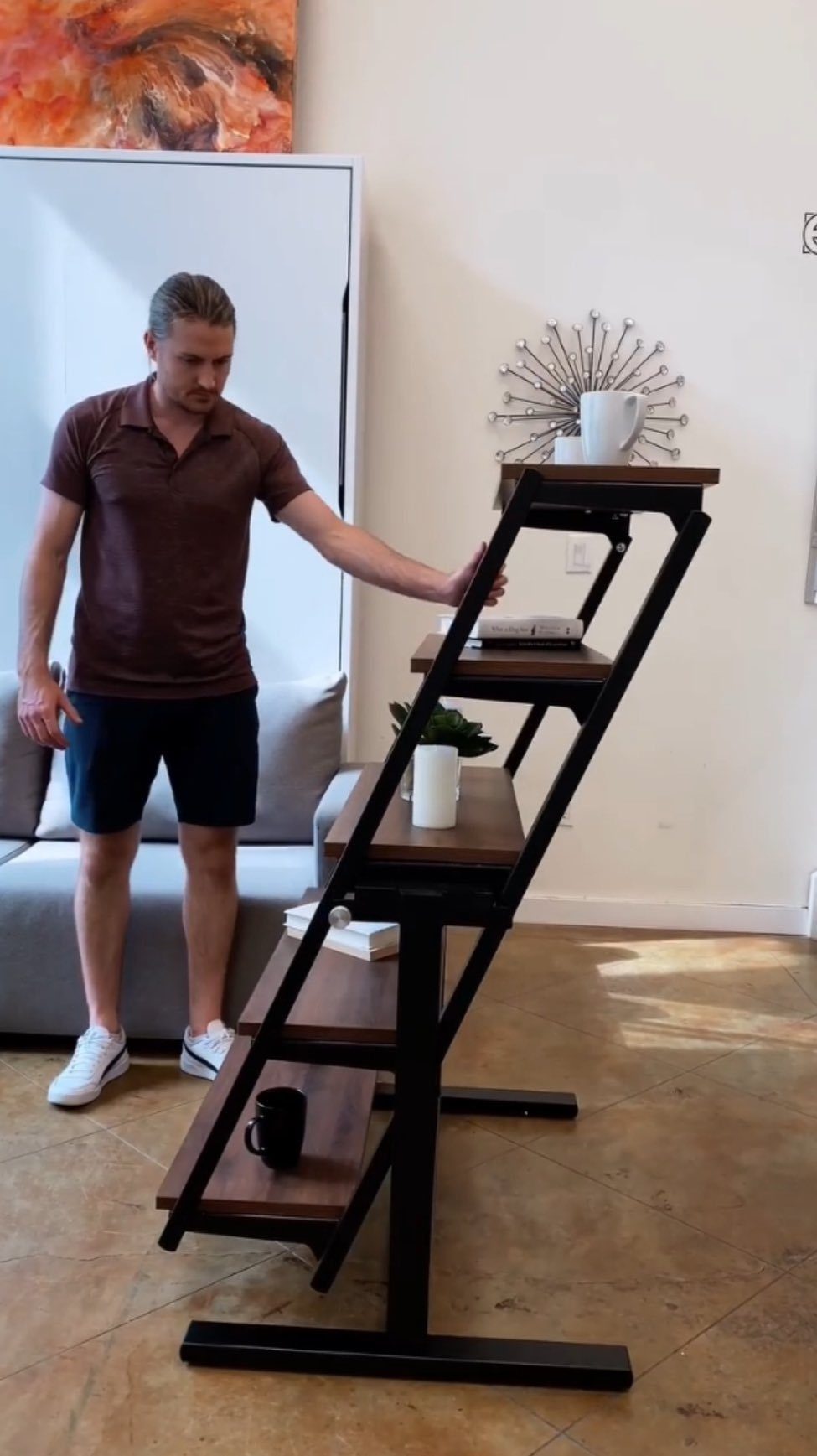 A man standing beside a dark wood and black metal leaning ladder shelf used as a multi-functional desk.