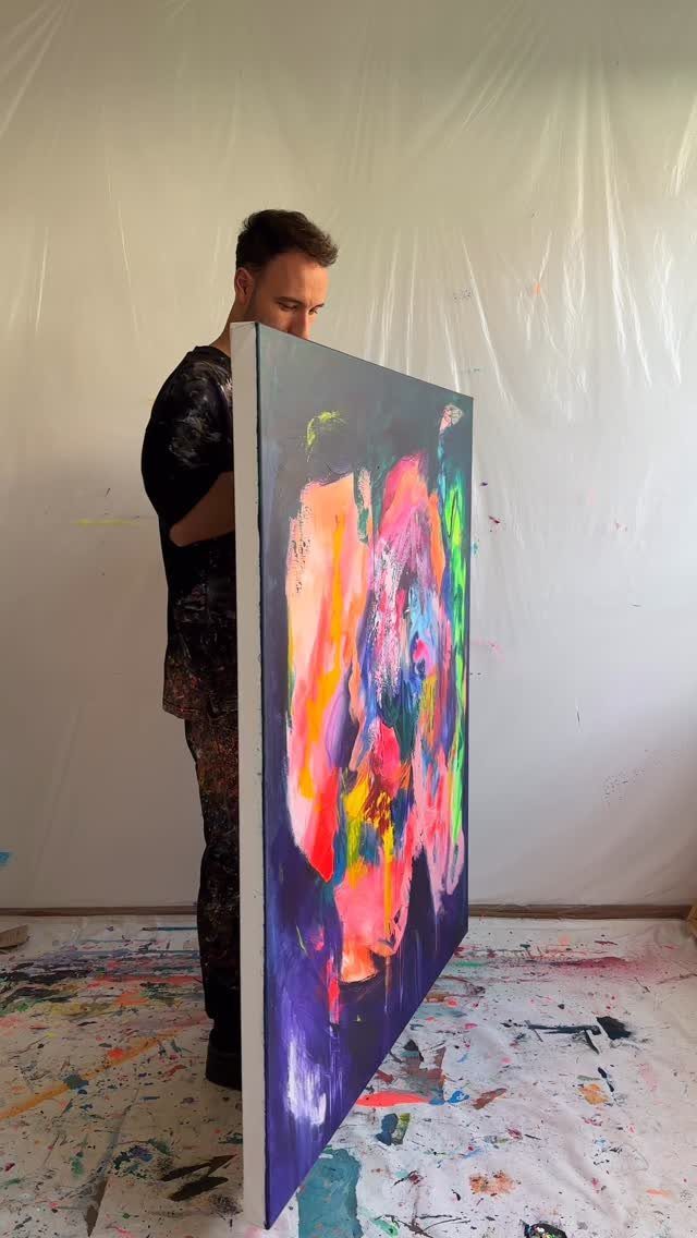A man holding a large, colorful abstract painting in a paint-splattered studio.