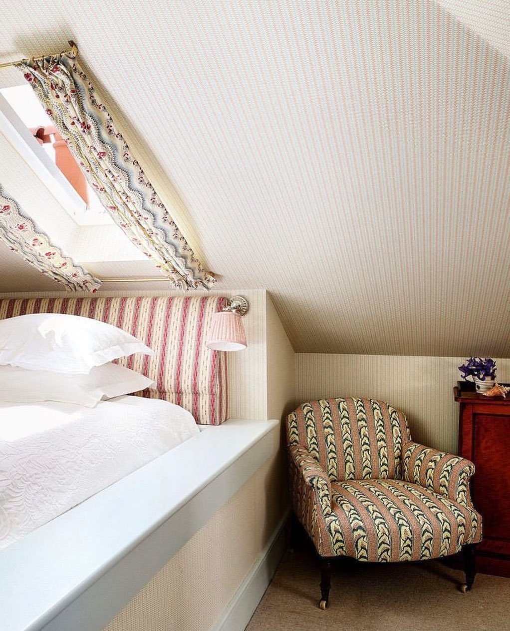 Sloped attic bedroom walls and ceiling covered in light patterned wallpaper near a bed.