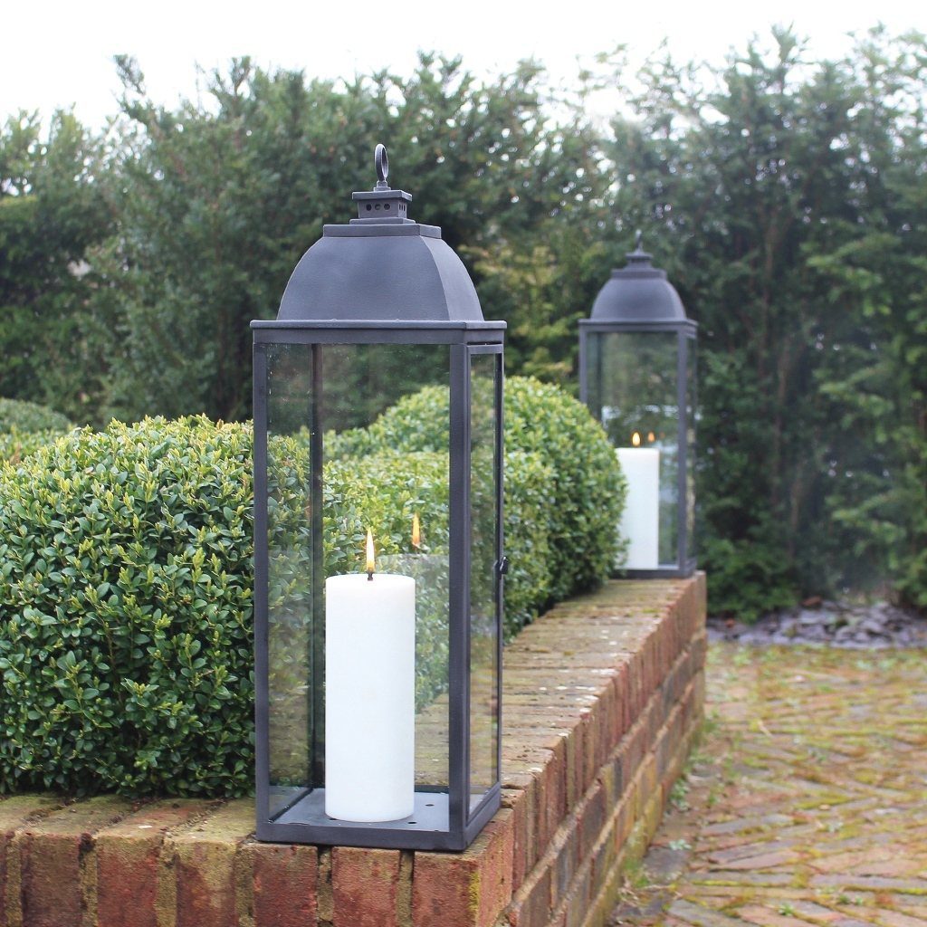Two large metal lanterns with lit white pillar candles resting on a brick wall.