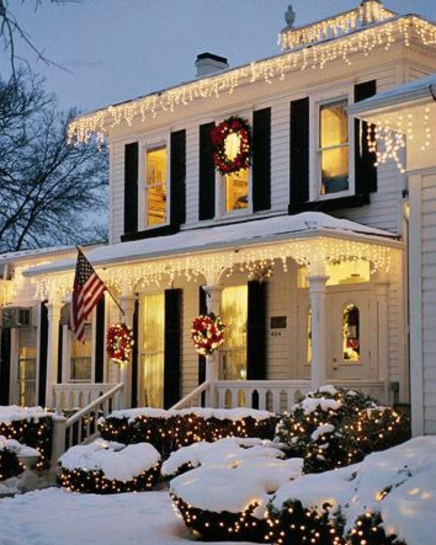 White house decorated with cascading icicle lights, red wreaths, and snow-covered bushes at twilight.