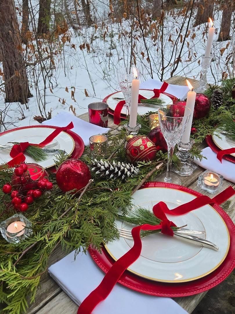 A festive outdoor table set with red plates, greenery, ornaments, and lit candles in a snowy forest.