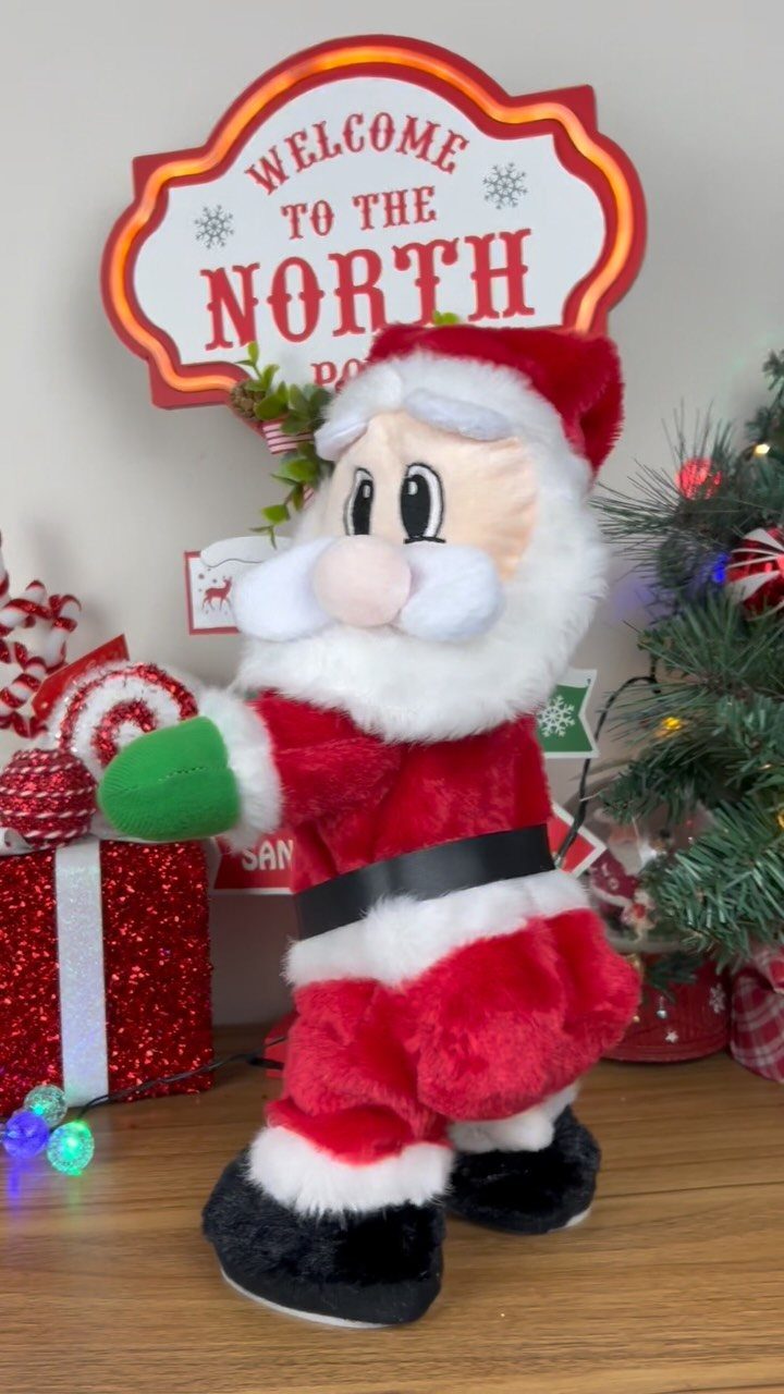 A large plush Santa Claus figure standing next to a North Pole sign and a small Christmas tree.