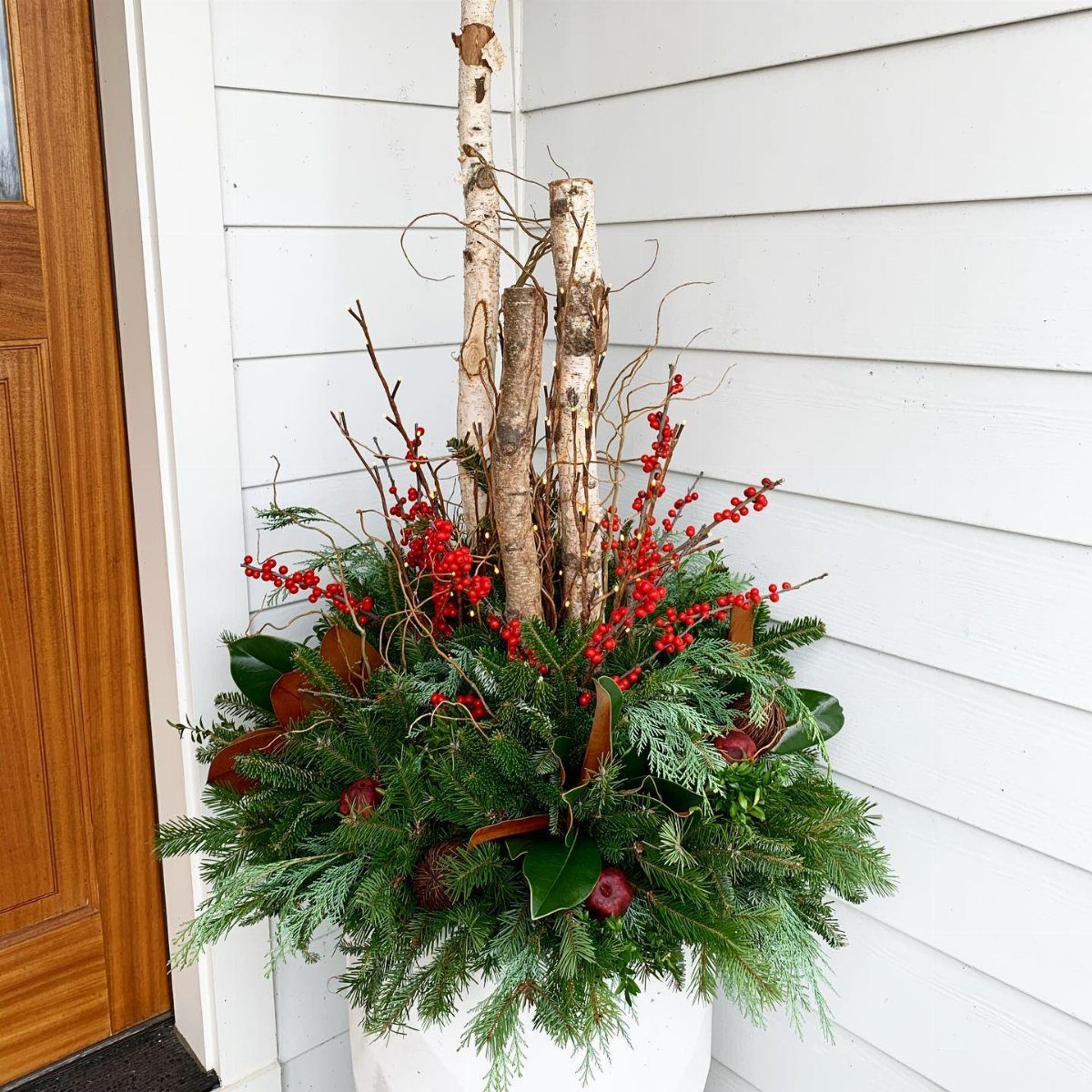 A white planter containing evergreen branches, red berries, and tall birch twigs against a white wall.