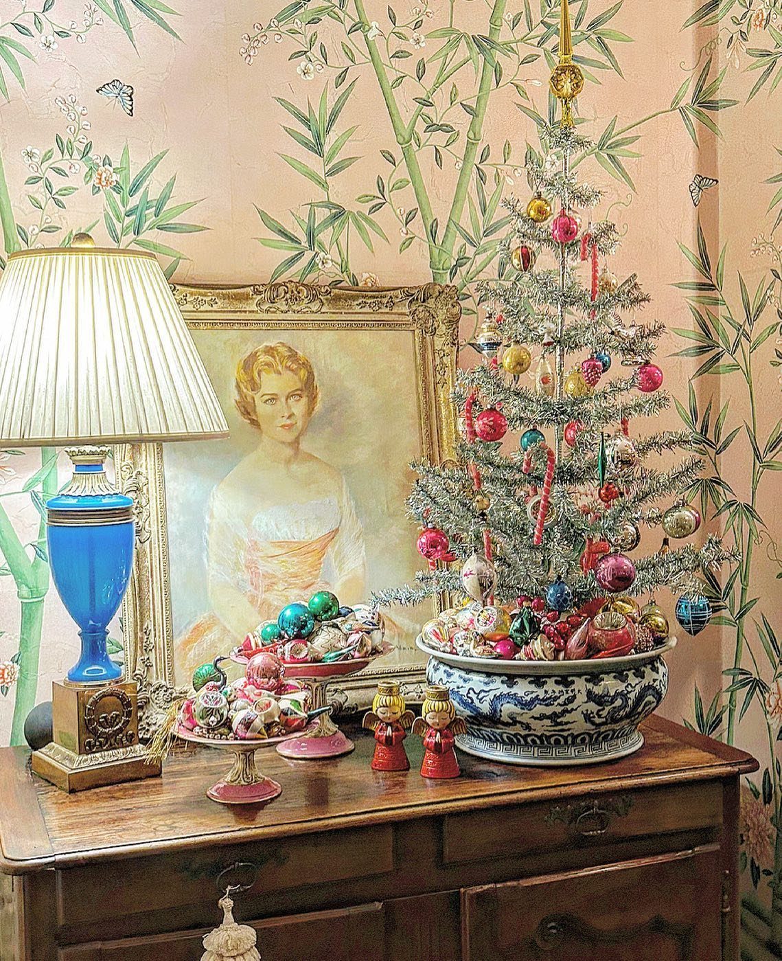 A silver tinsel Christmas tree with colorful ornaments sits on a wooden sideboard next to a lamp.