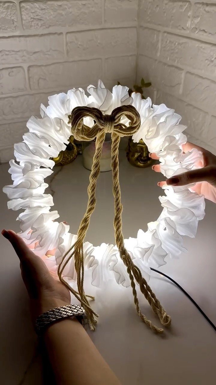 Hands holding a glowing white ruffled fabric wreath with a large jute rope bow.