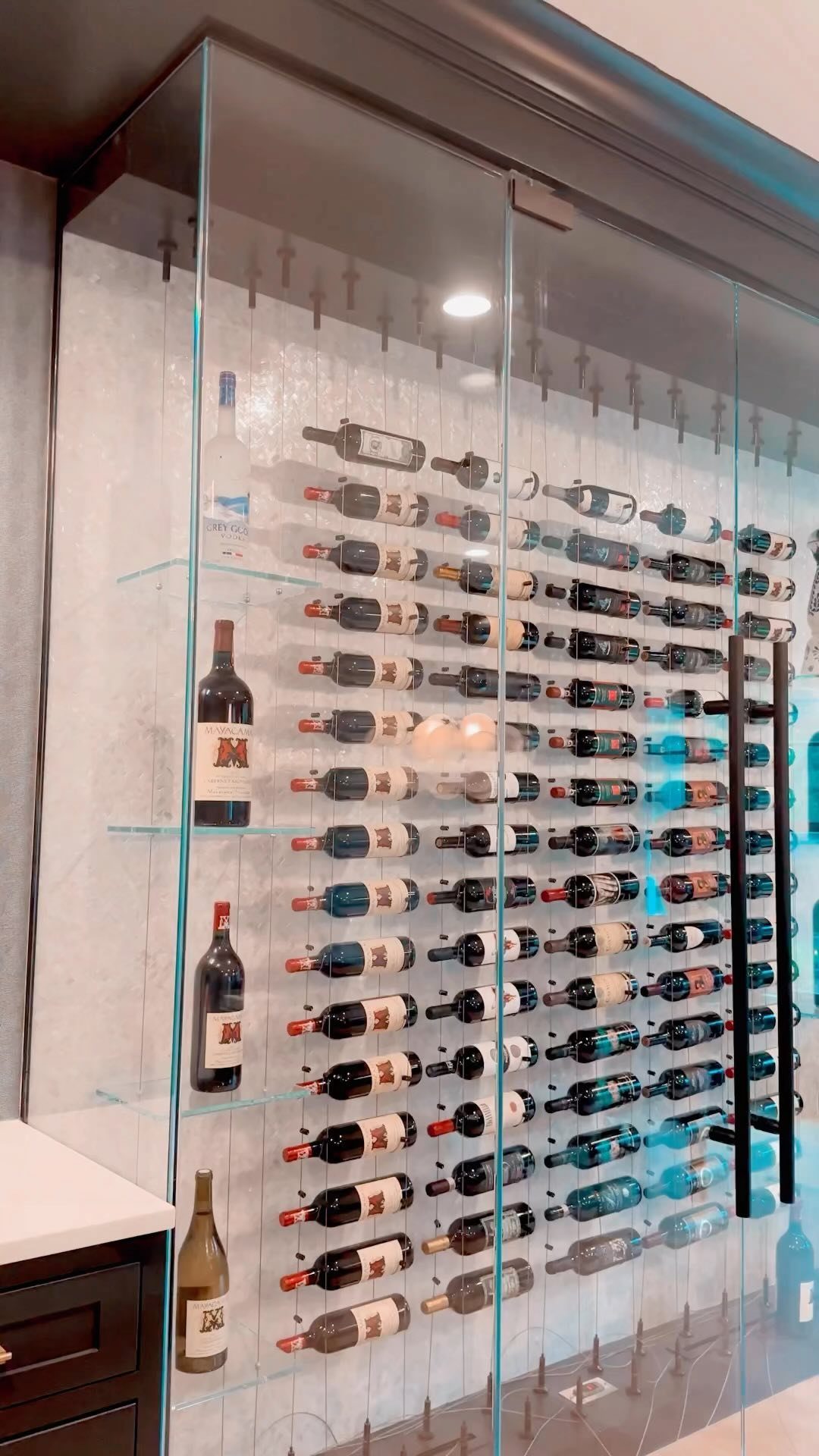 Wine bottles stored horizontally on metal racks inside a glass-enclosed cellar with blue lighting.