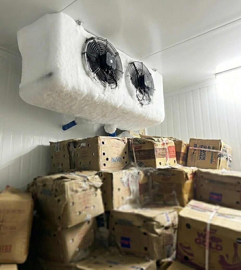 Industrial refrigeration unit mounted above stacks of cardboard produce boxes in a cold storage room.