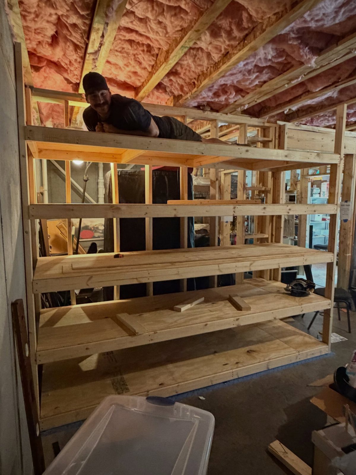 A man leaning over large, newly built wooden shelving units in a basement with pink insulation.