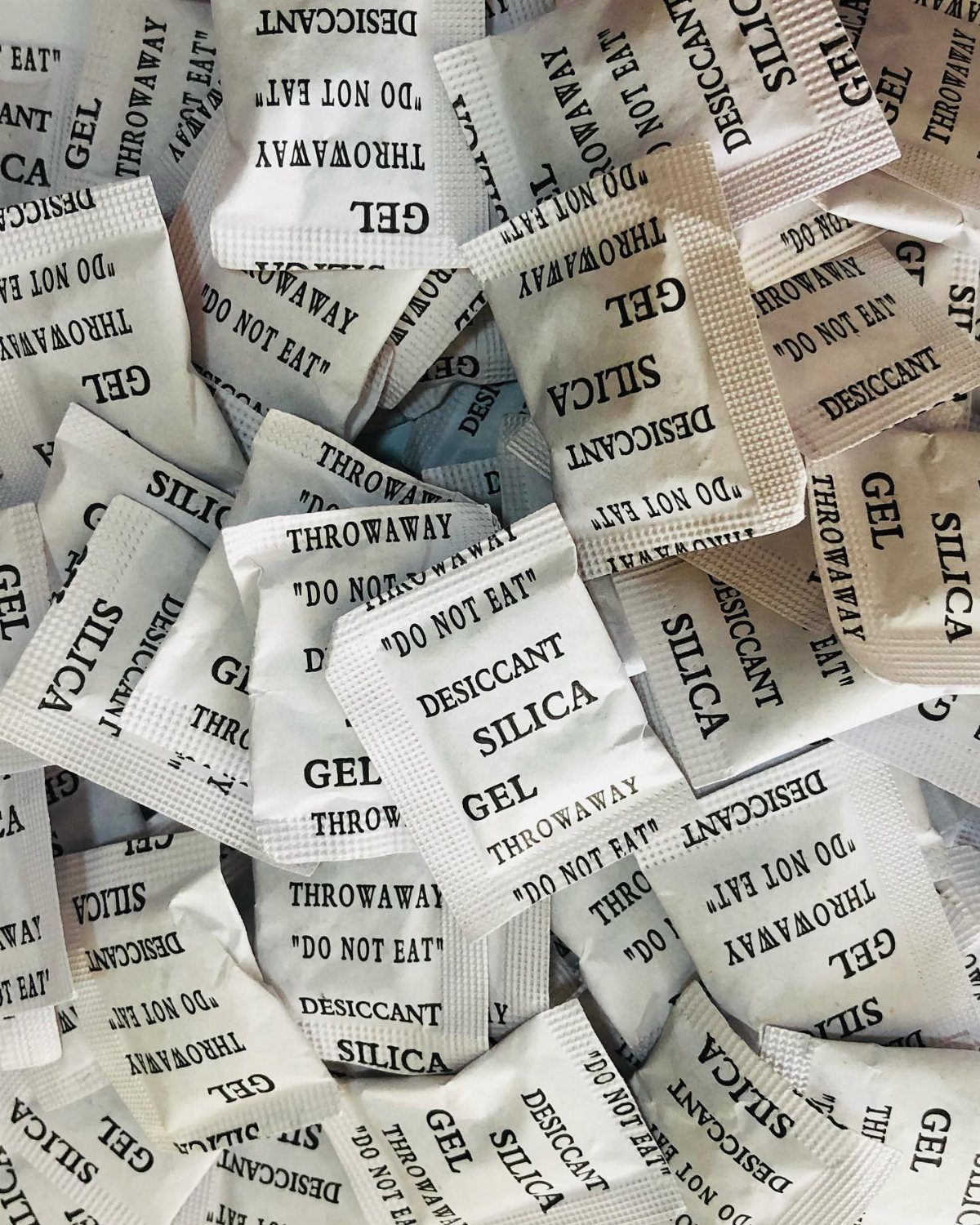 Numerous white silica gel desiccant packets piled together with black printed warning text.