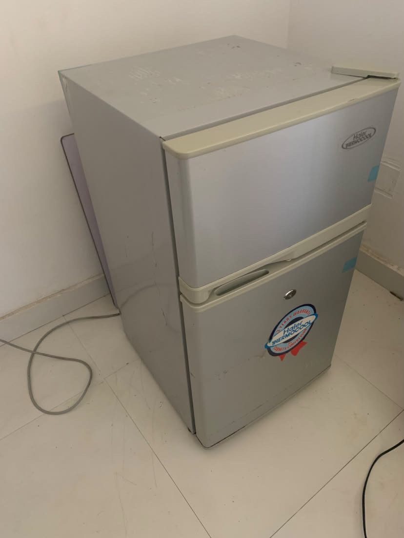 A small silver two-door compact refrigerator standing in a corner on a tiled floor.