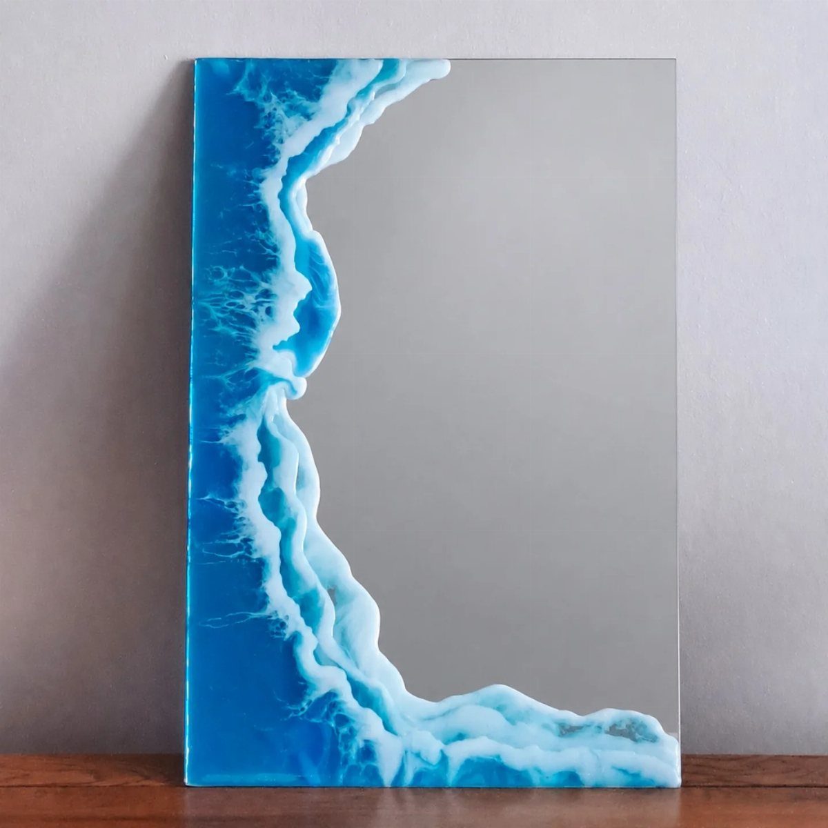 Ocean wave resin art mirror leaning against a grey wall on a wooden surface.
