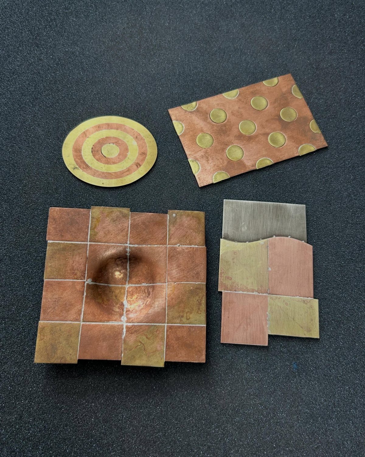 Metal tiles and a circular disc featuring copper, brass, and silver finishes on a dark surface.