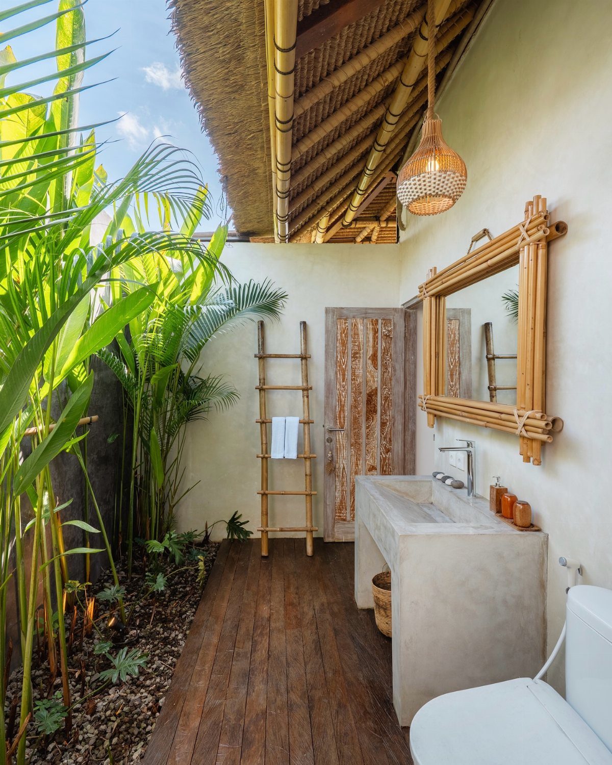 Outdoor bathroom with concrete sink, wooden floors, bamboo mirror, and tropical greenery under a thatched roof.