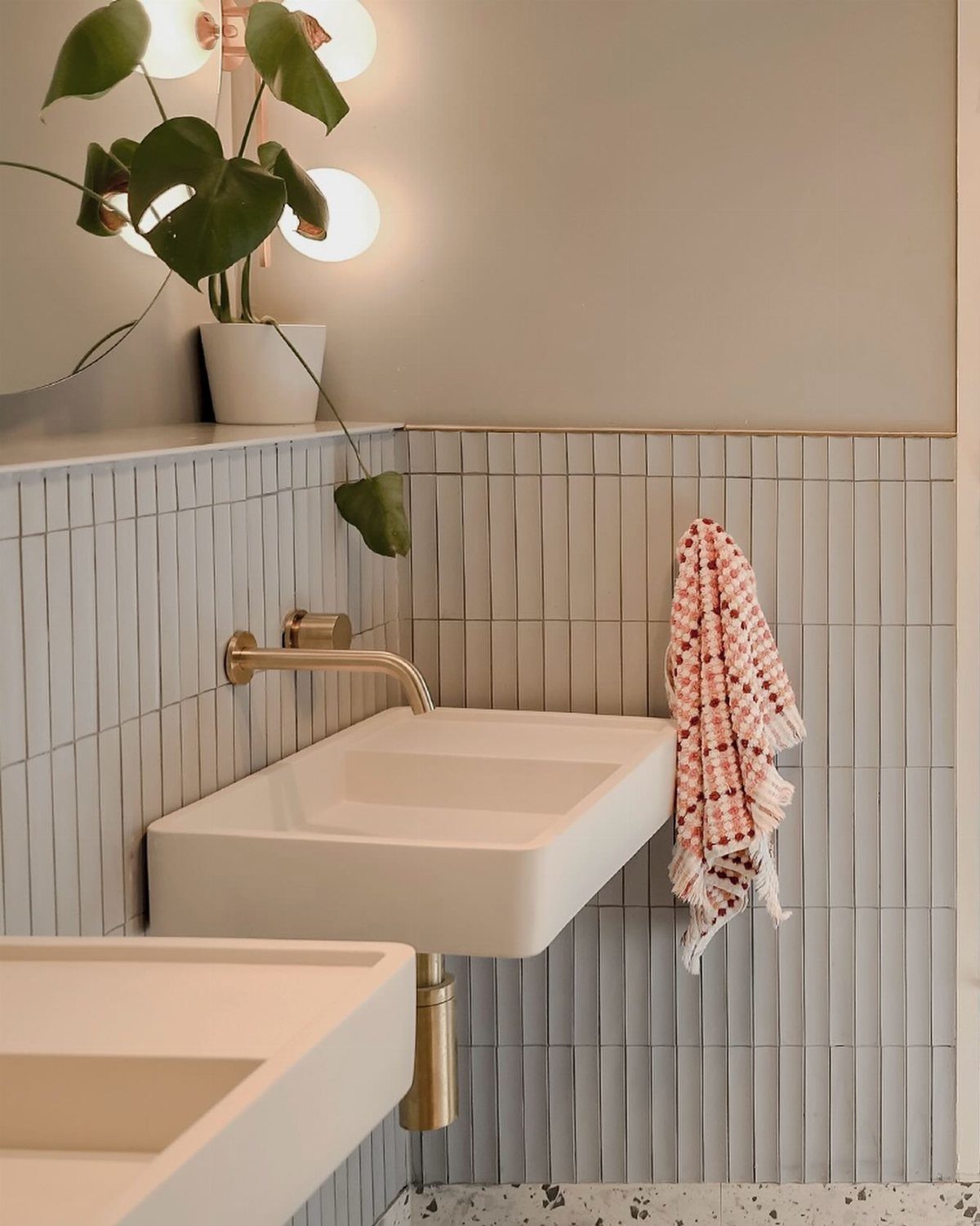 A white wall-mounted sink with gold hardware and a small potted plant on a shelf.