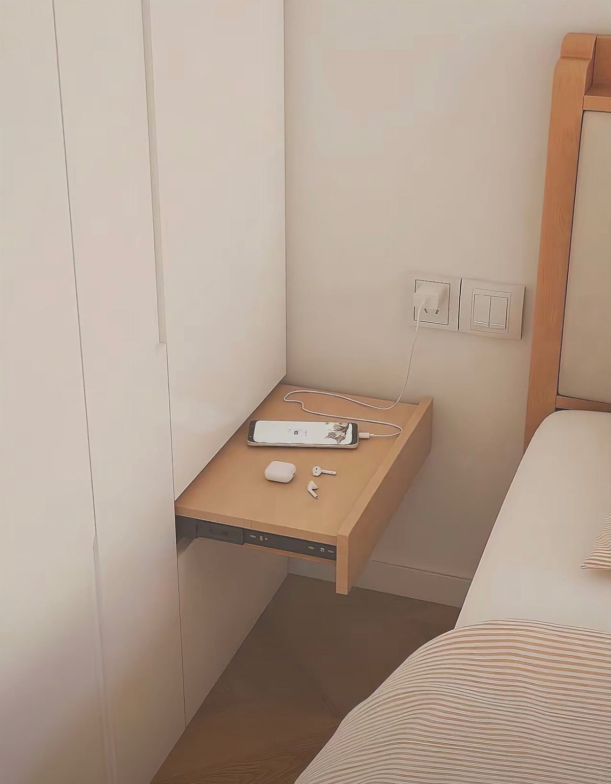 Light wood floating corner shelf holding a smartphone and earbuds next to a bed.