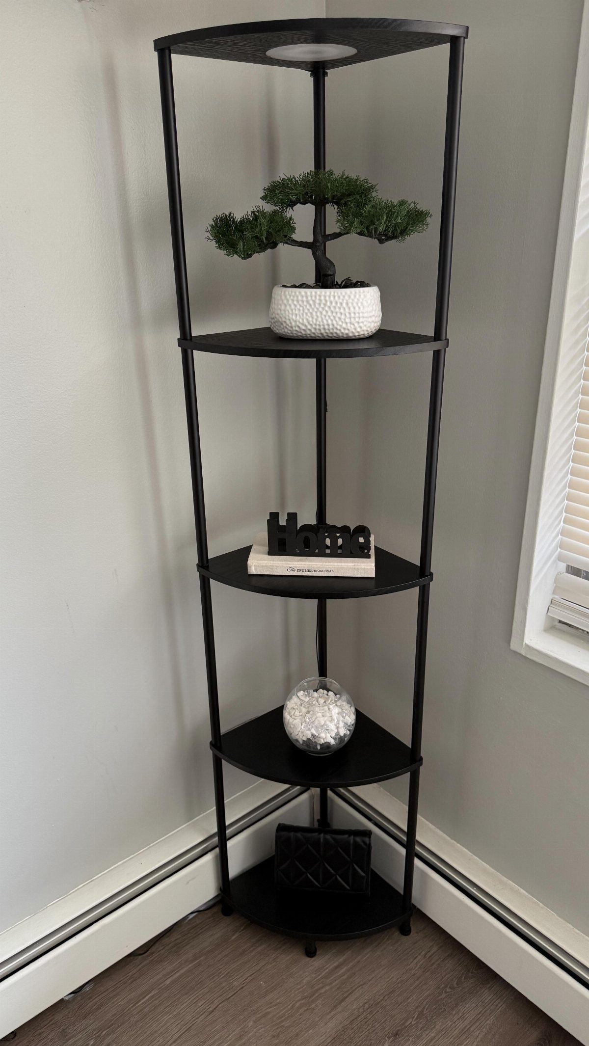 Black metal corner shelf unit holding a bonsai tree, books, and decorative glass bowl.