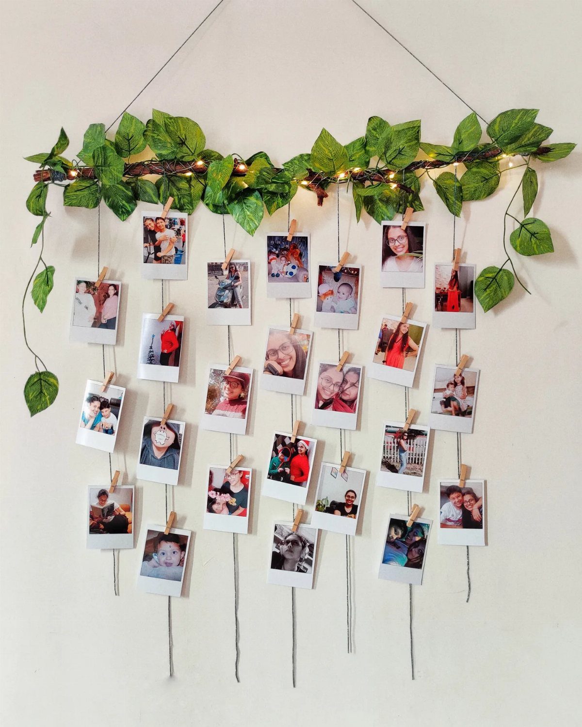 Polaroid photos hanging from a leafy vine with fairy lights on a white wall.