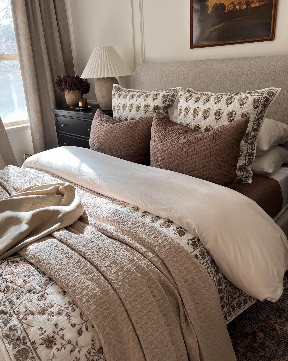 A cozy bed with layered bedding, patterned pillows, and a lamp on a nightstand.