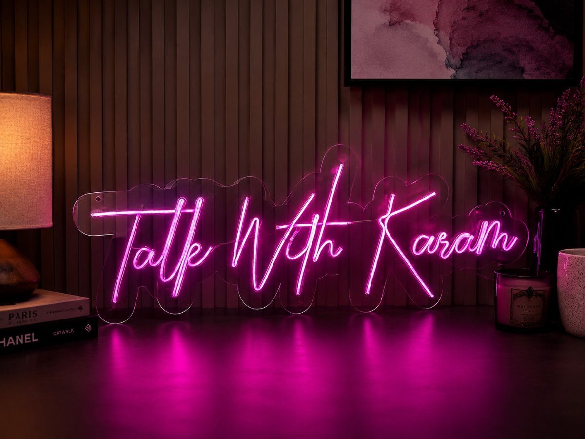Pink neon sign reading Talk With Karam on a dark tabletop near a lamp.