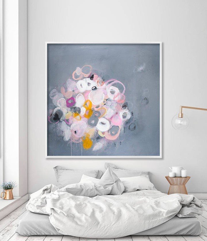 Abstract colorful painting in a white frame hanging on a white wall above a bed.