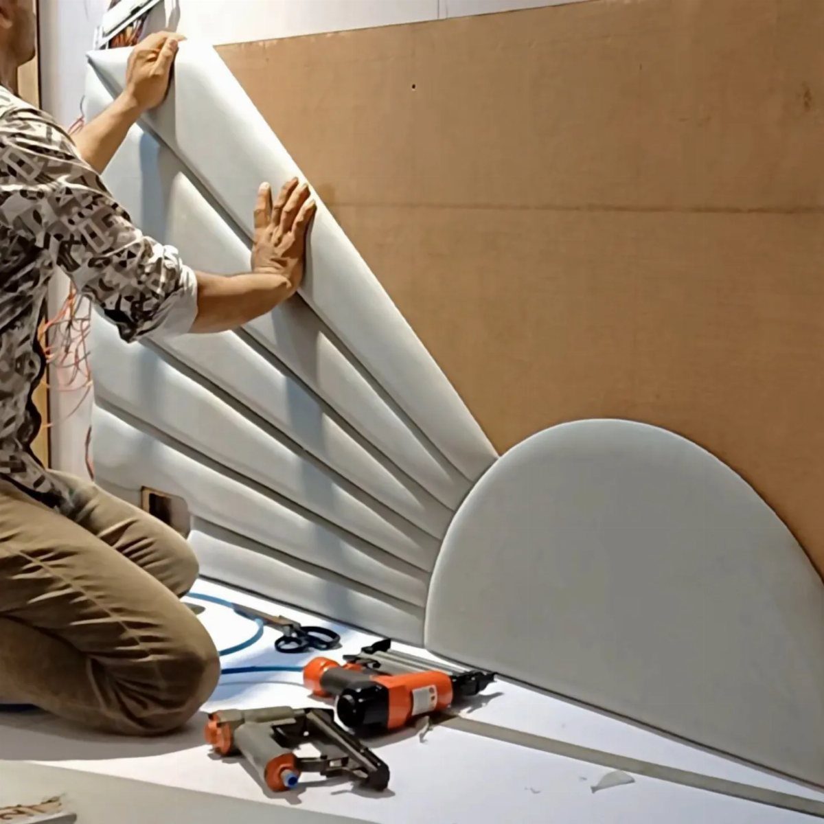 A person installing large grey upholstered curved panels onto a wooden wall as a DIY headboard.
