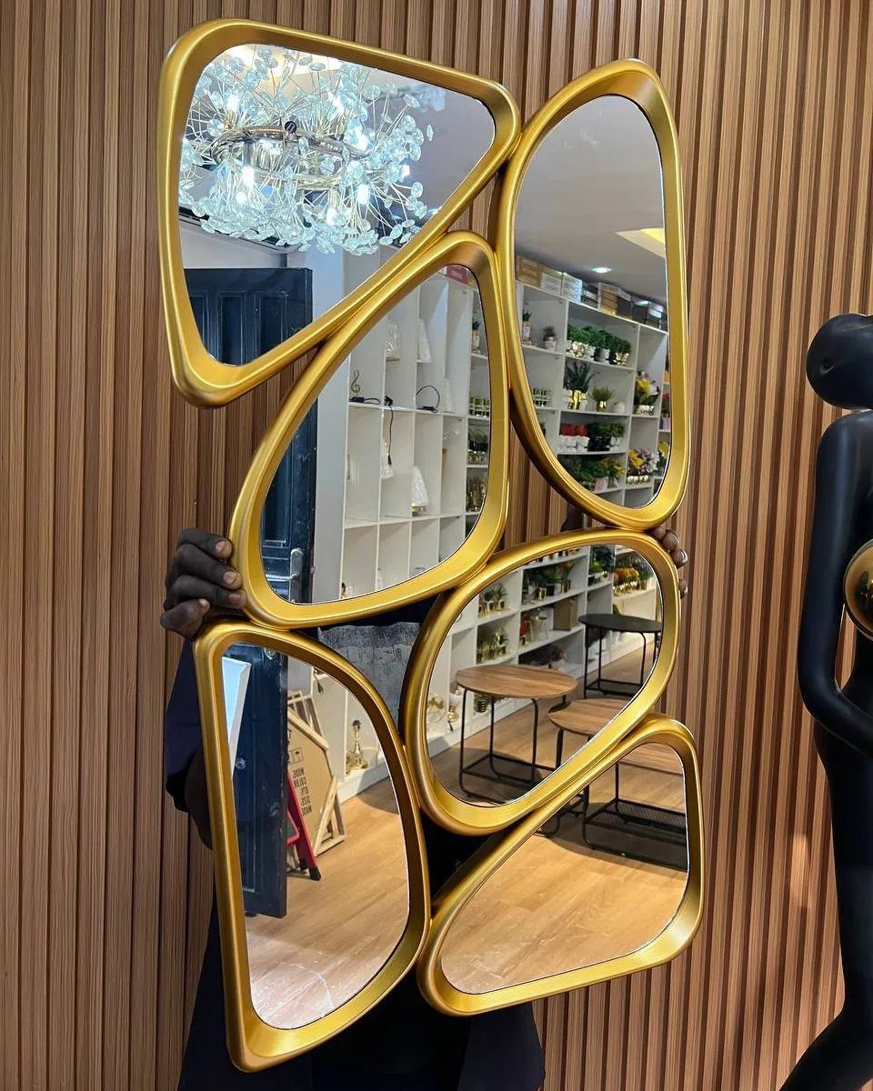 A person holding a large, organic-shaped gold multi-paneled mirror against a wood-slat wall.