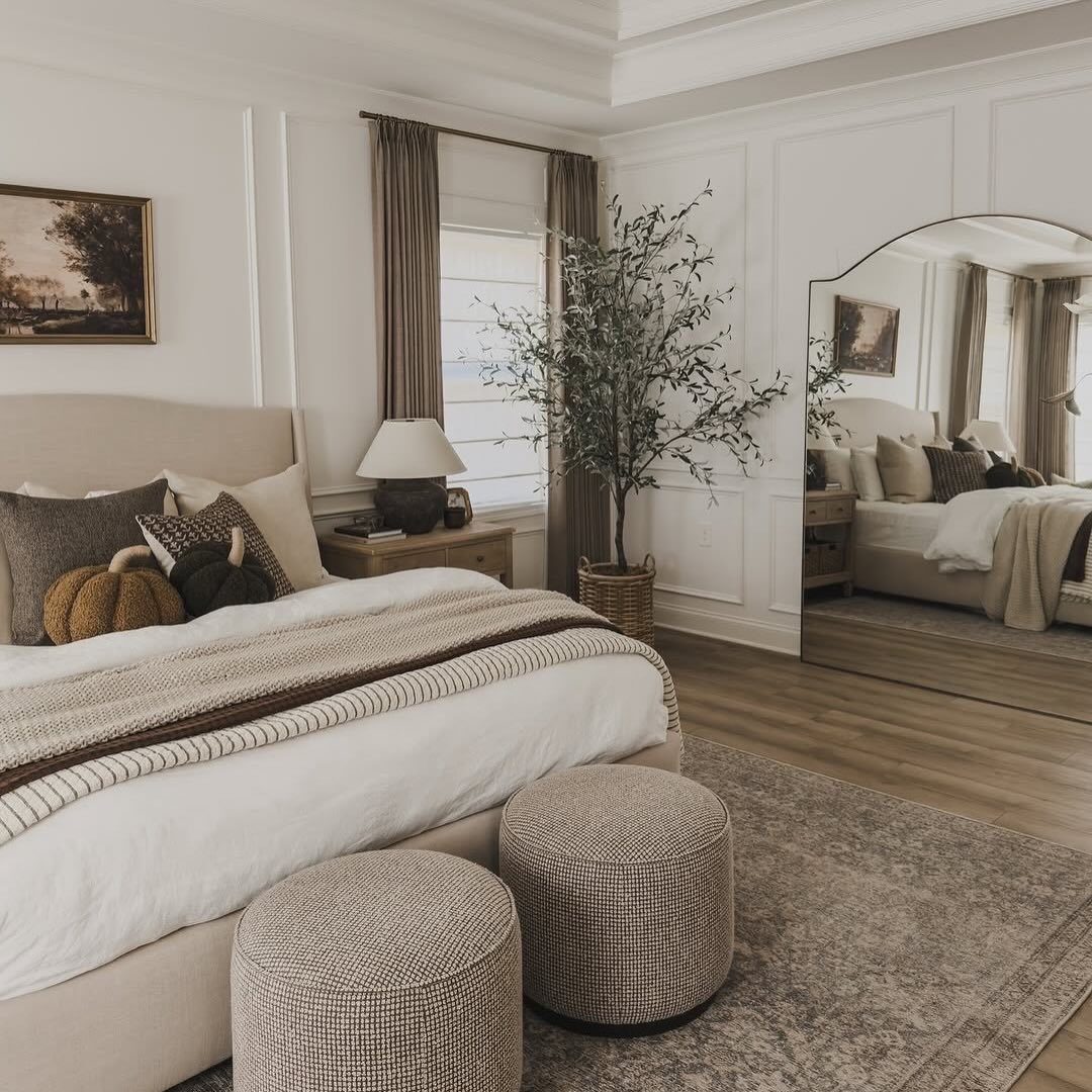 Neutral bedroom with white paneled walls, a large bed, textured ottomans, and an arched mirror.