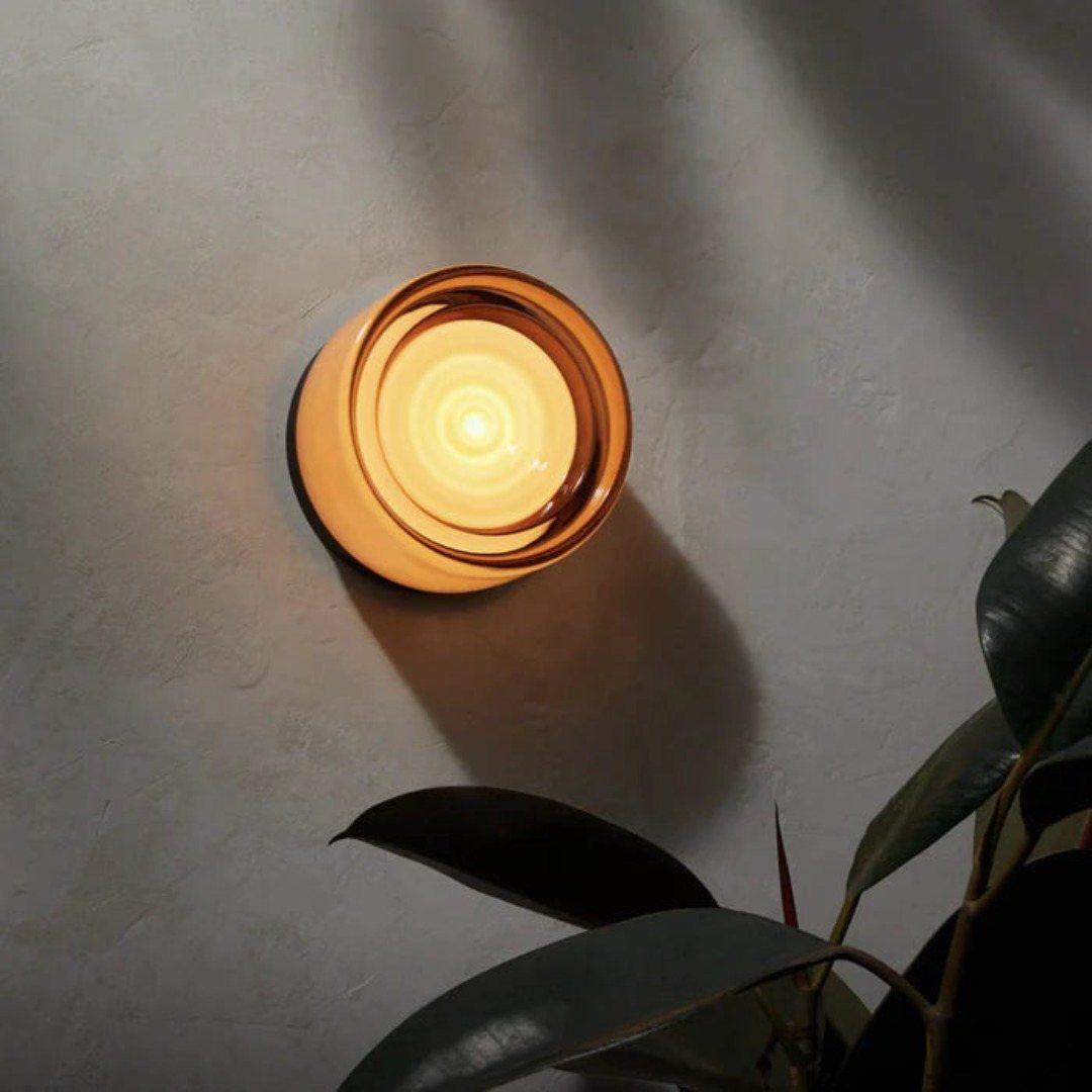 A glowing amber glass wall sconce mounted on a textured grey wall near plant leaves.