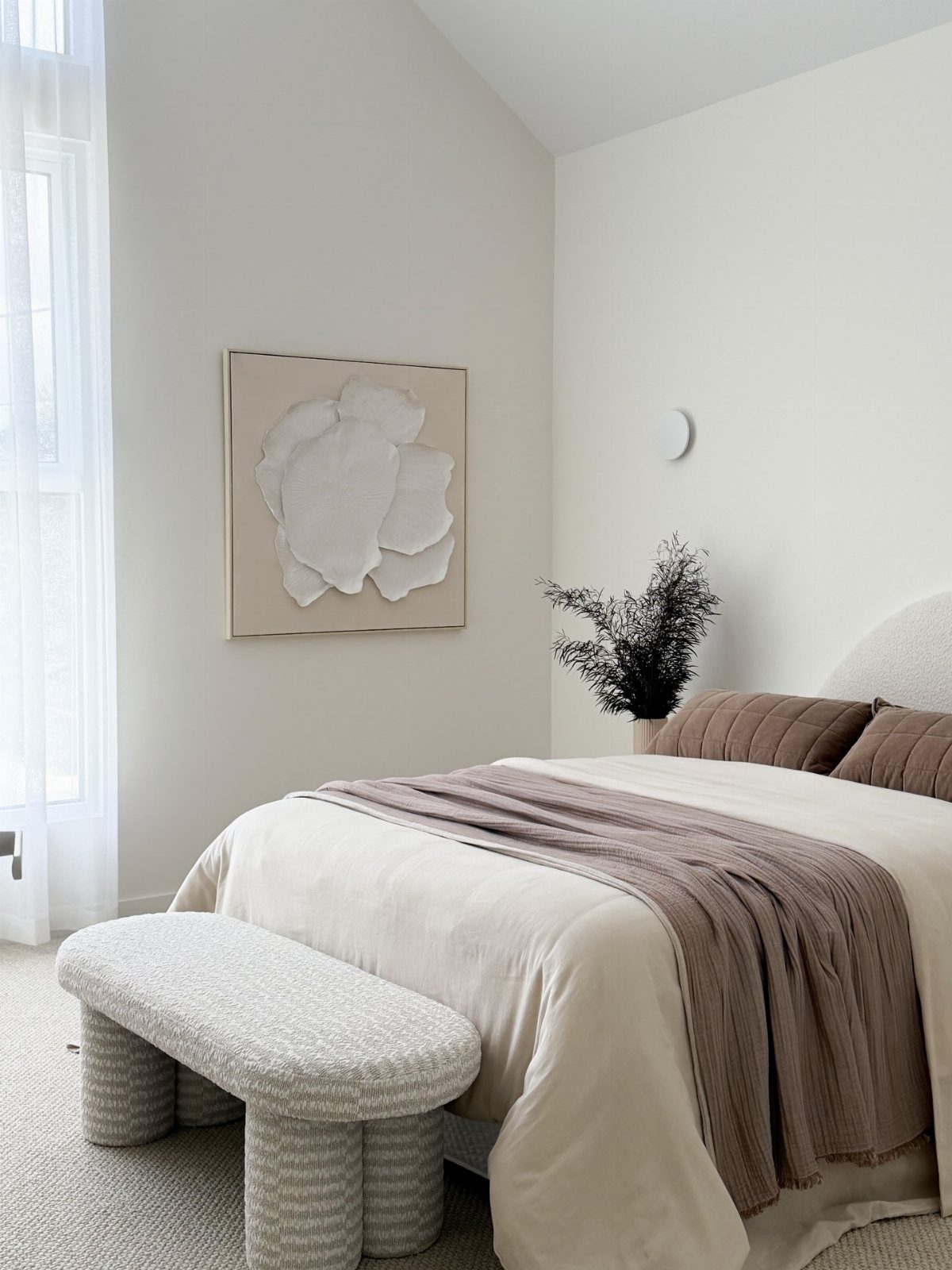 A minimalist bedroom with cream bedding, a mauve throw, and a textured grey bench.