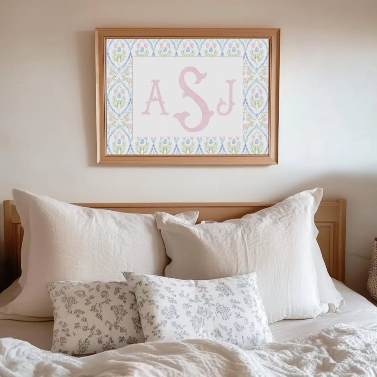 Framed pink monogram art hanging above a bed with white and floral pillows.