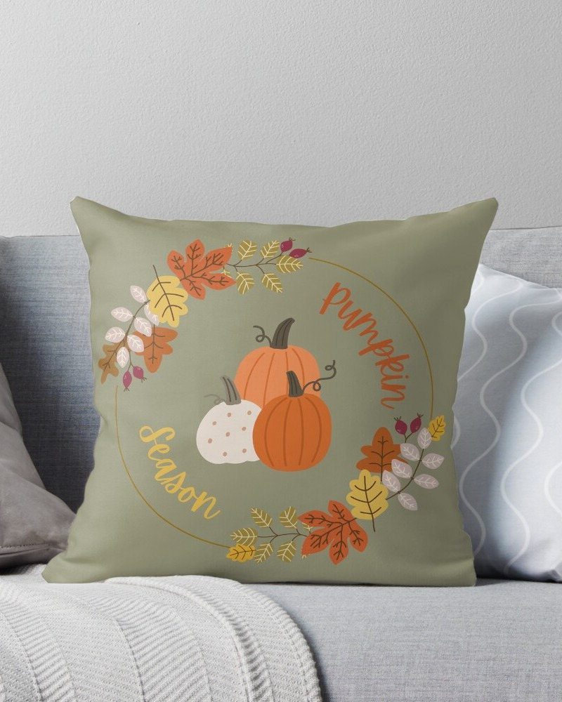 Sage green throw pillow with pumpkin and autumn leaf illustrations resting on a grey sofa.