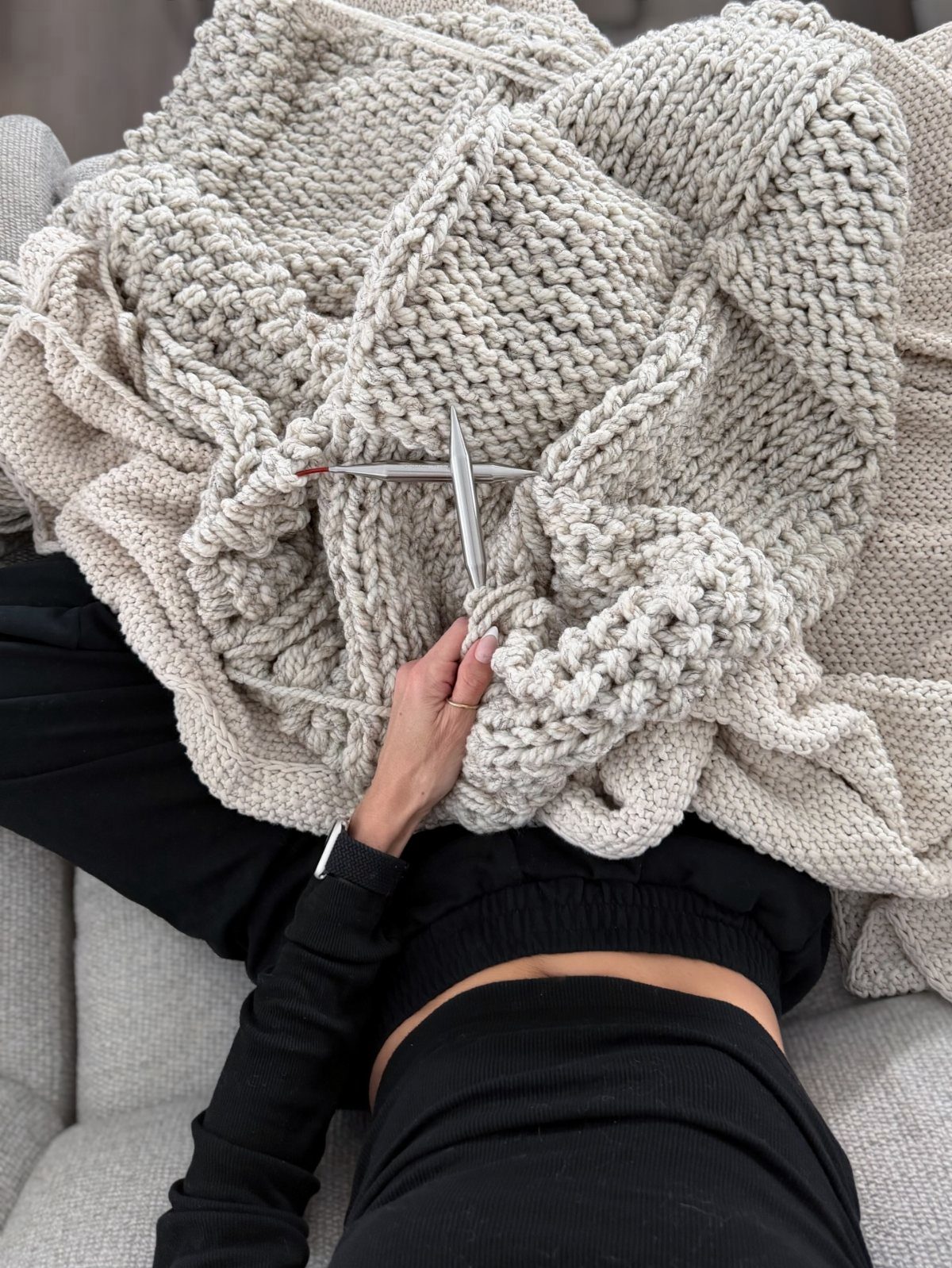 Hands knitting a large cream chunky knit blanket with silver needles.