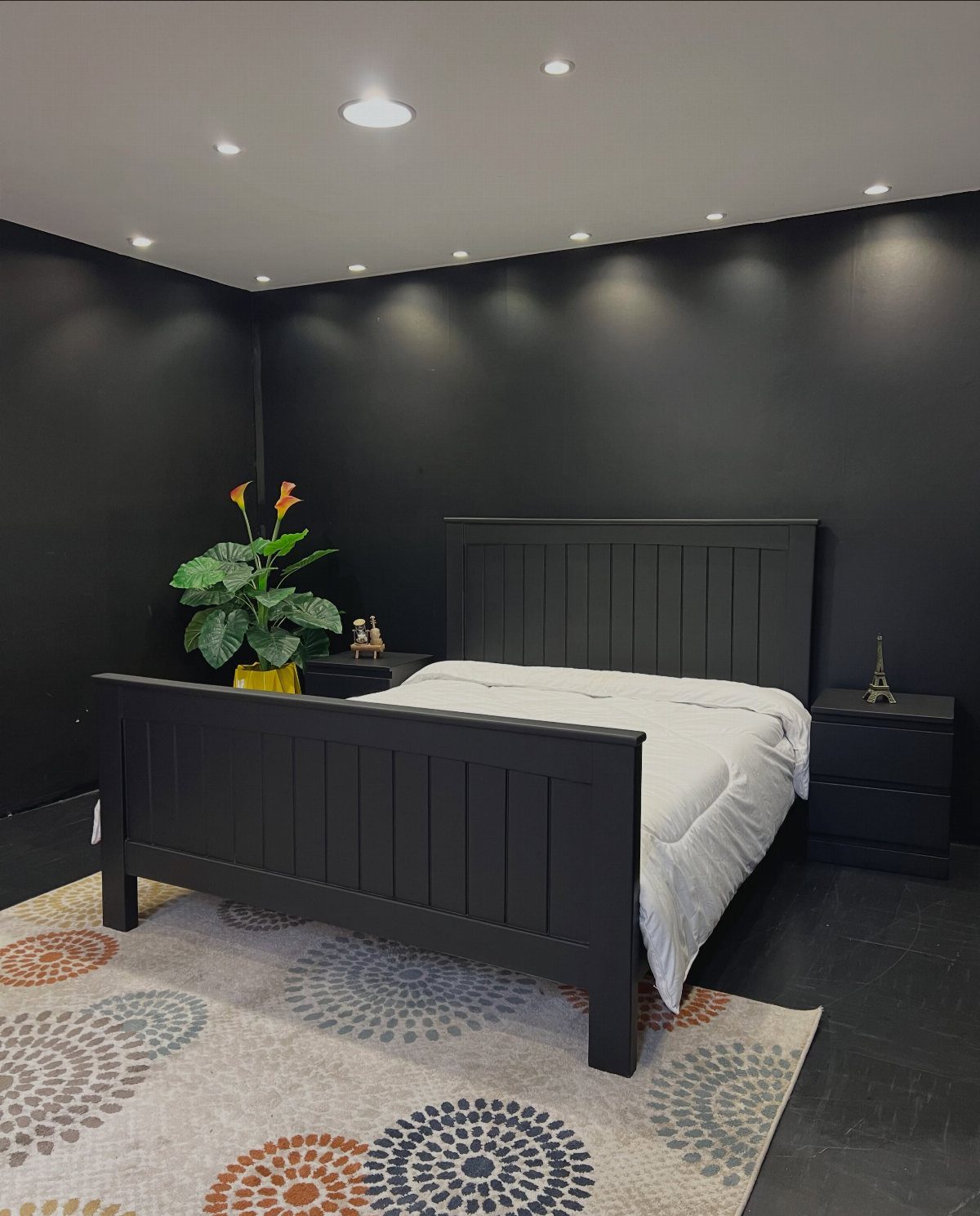 Black wooden bed frame with white bedding in a room with black walls and a patterned rug.