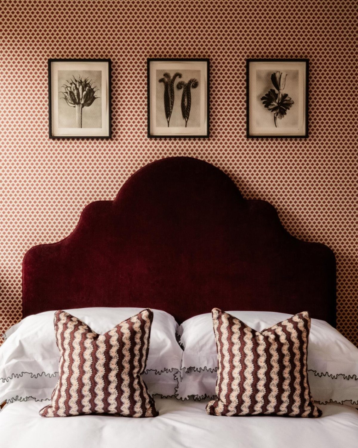 Burgundy velvet scalloped headboard against patterned wallpaper with botanical art and patterned pillows.