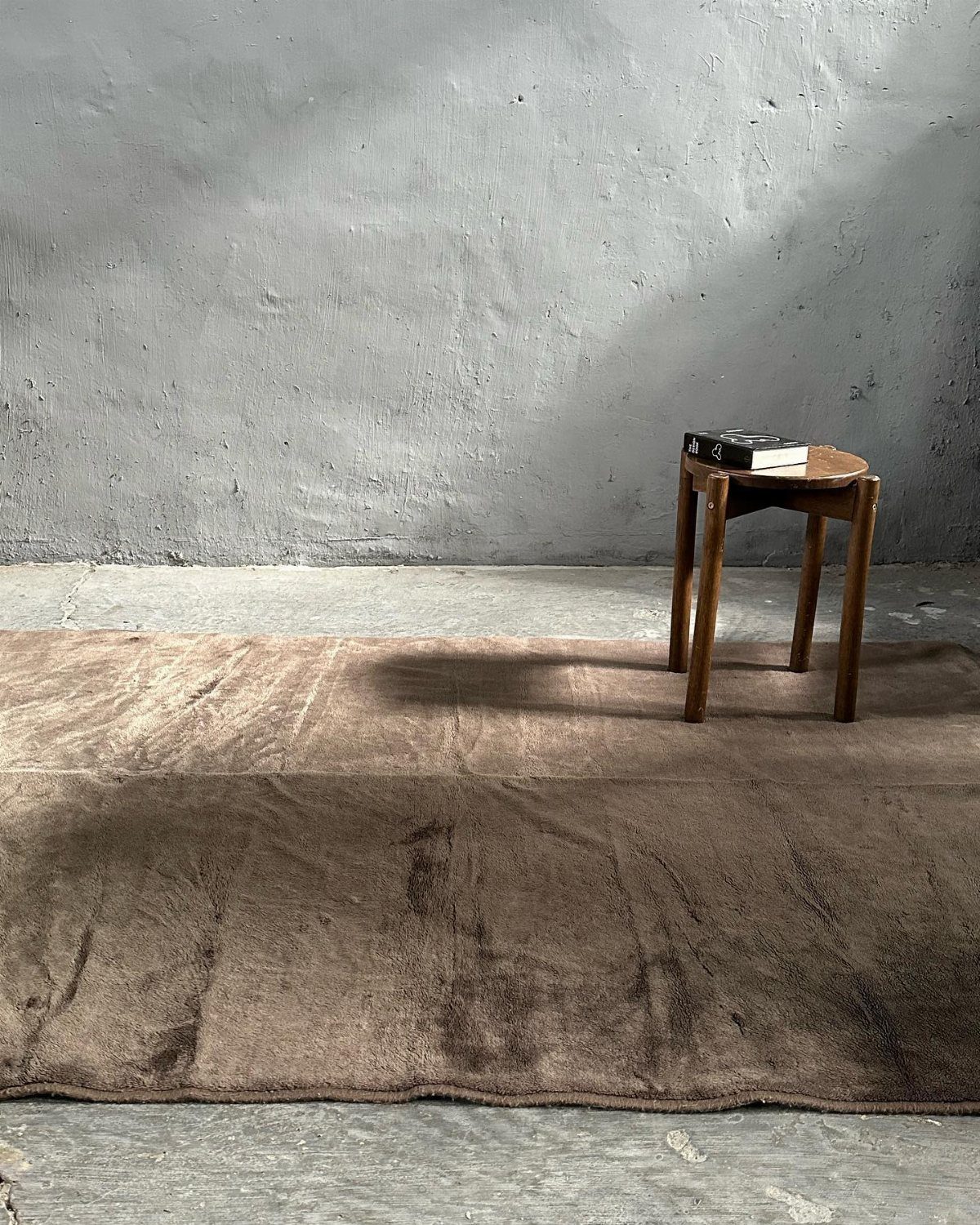 A large textured brown area rug on a concrete floor next to a small wooden stool.