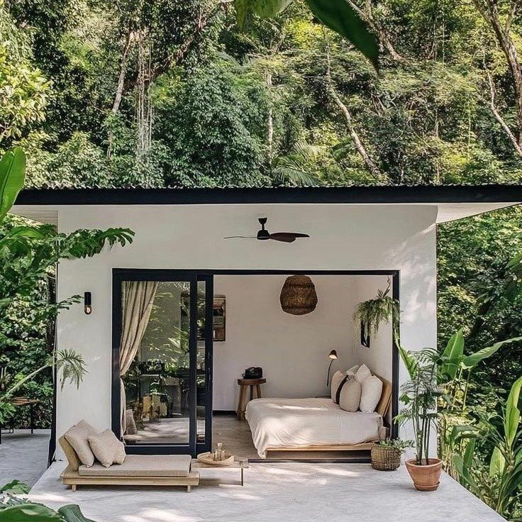 Minimalist white cabin bedroom with light bedding and wooden furniture surrounded by dense green forest.
