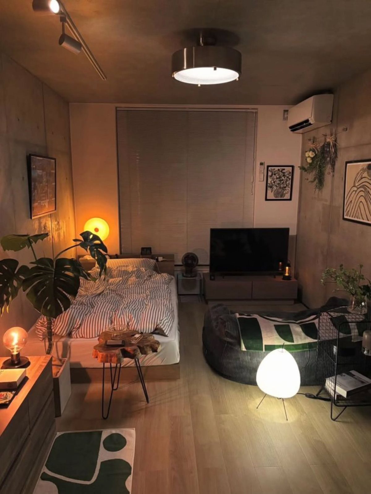 A cozy bedroom with warm orb lighting, striped bedding, and green plants against concrete walls.