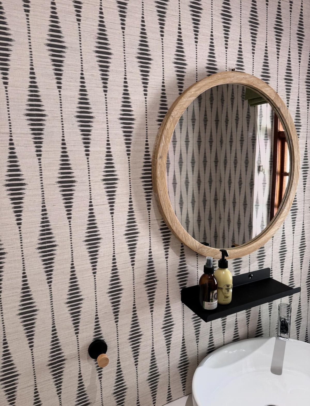 Black and cream patterned wallpaper on a bathroom wall with a round wooden mirror.