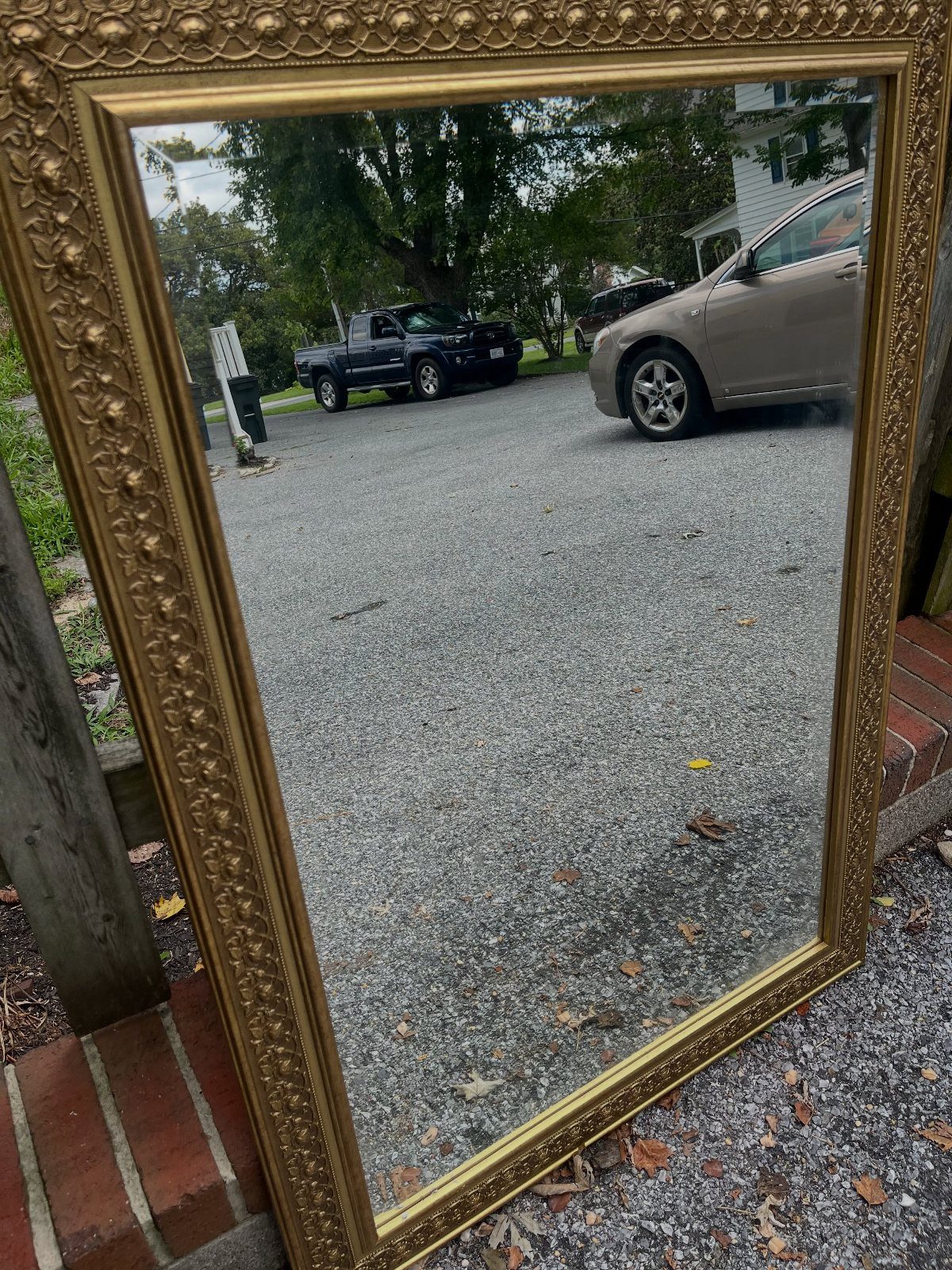 Ornate gold-framed mirror leaning outdoors reflecting a driveway with cars and trees.