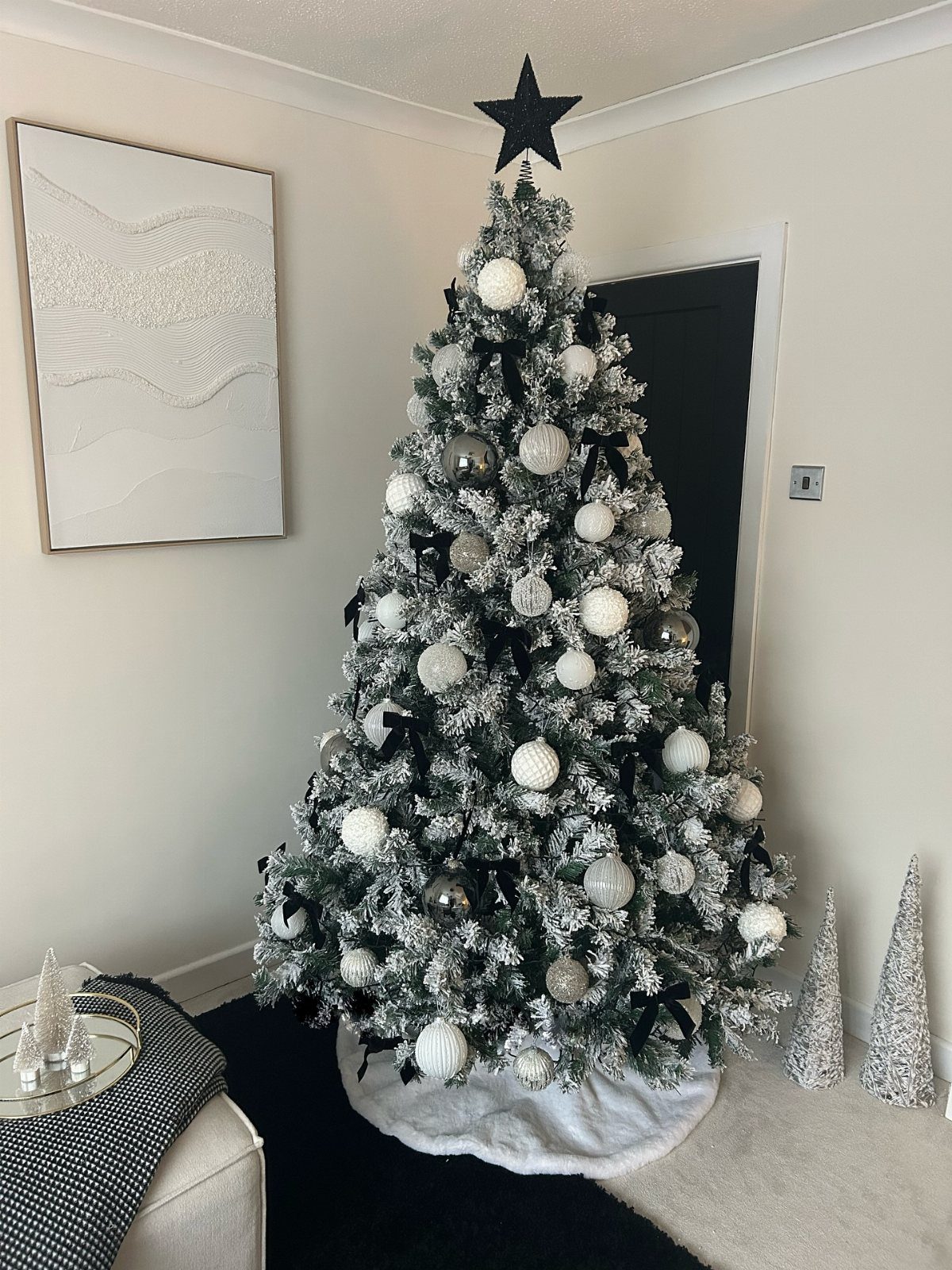 A flocked Christmas tree decorated with white ornaments and black ribbons against a neutral room.