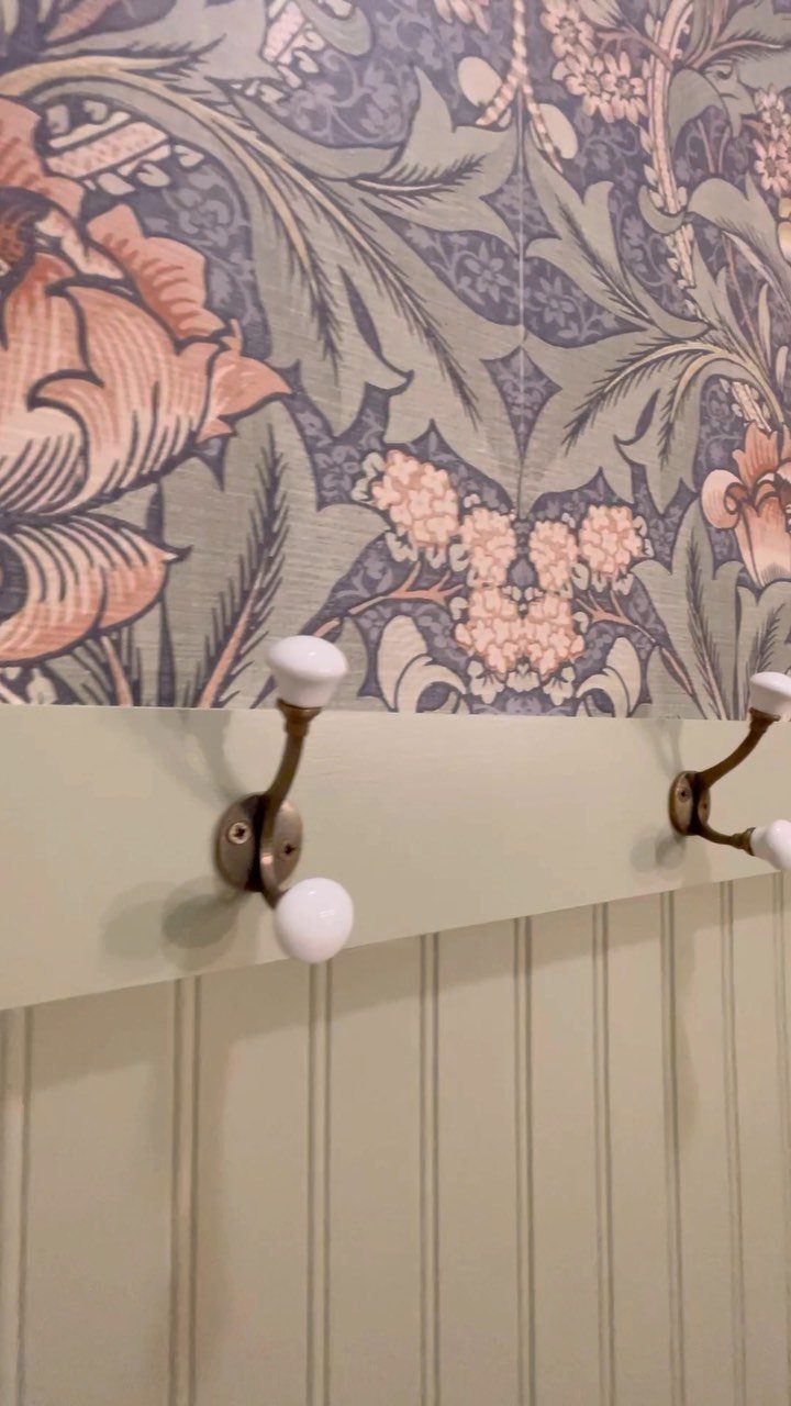 Floral patterned wallpaper above sage green beadboard with two brass wall hooks.