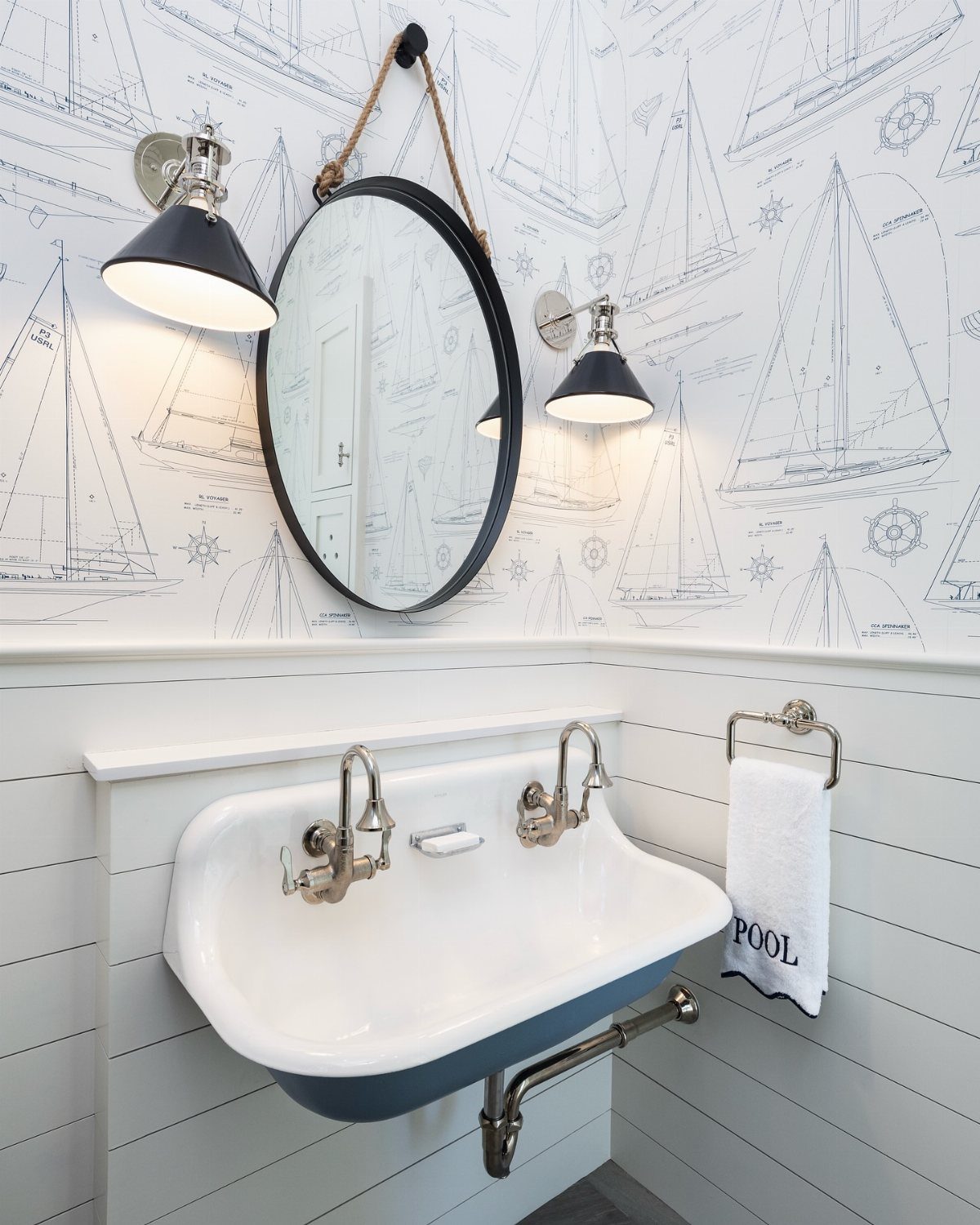 White bathroom with sailboat wallpaper, a blue sink, and a round mirror hanging by a rope.