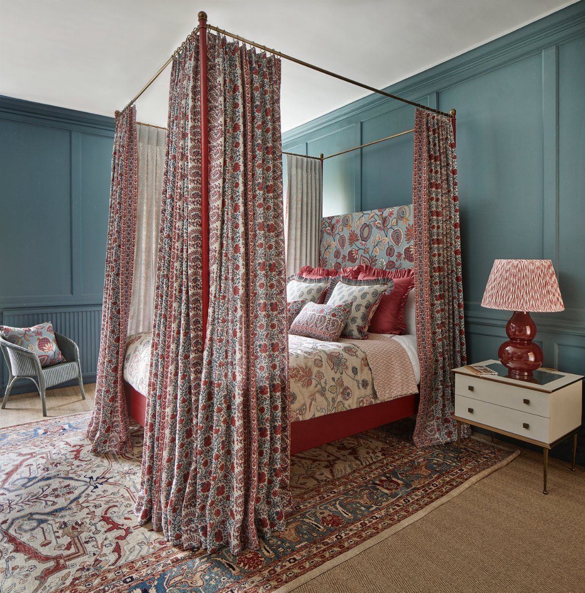 A canopy bed with floral patterned curtains and layered bedding in a blue paneled bedroom.