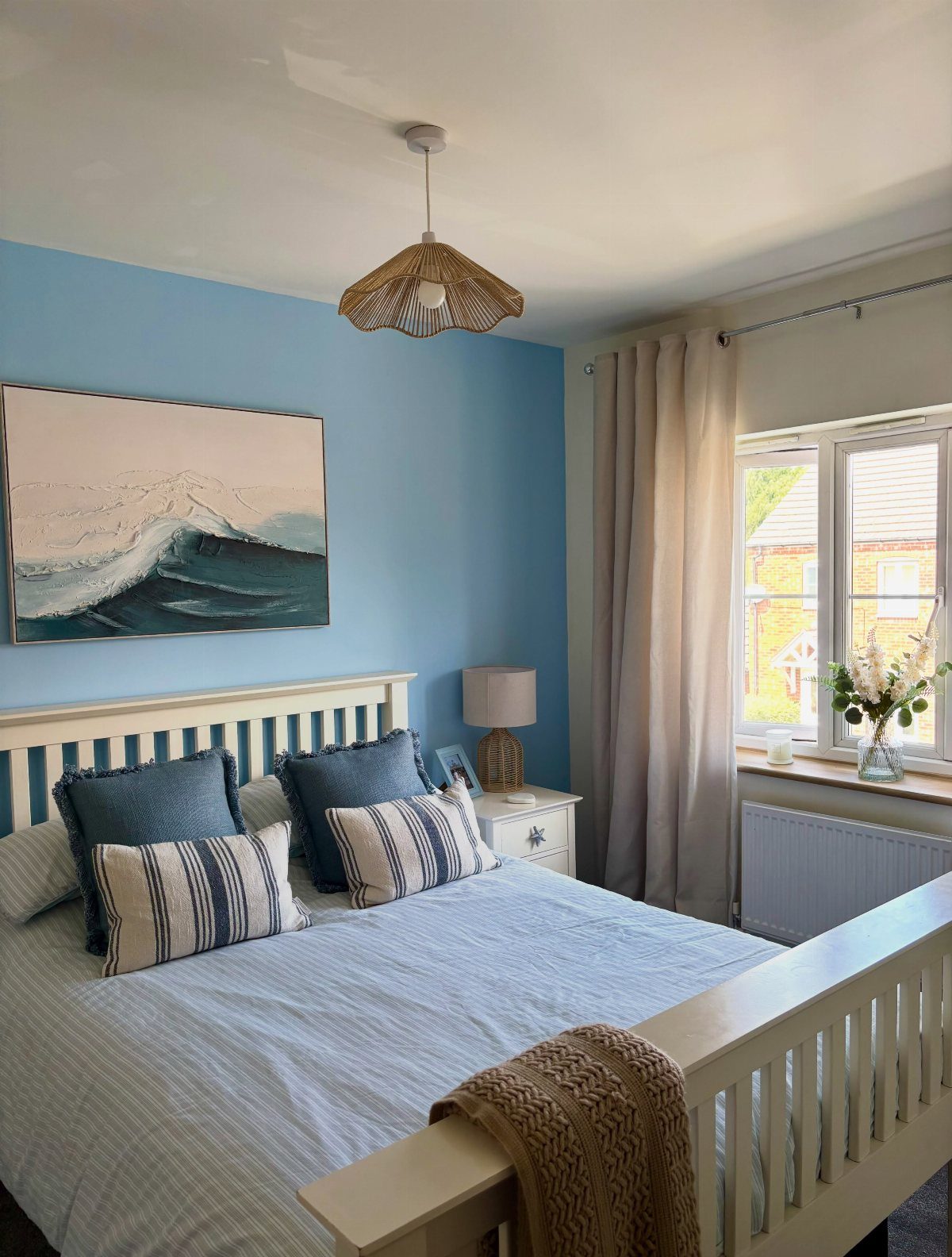 Light blue bedroom with white wooden bed, patterned pillows, and a wave painting on the wall.