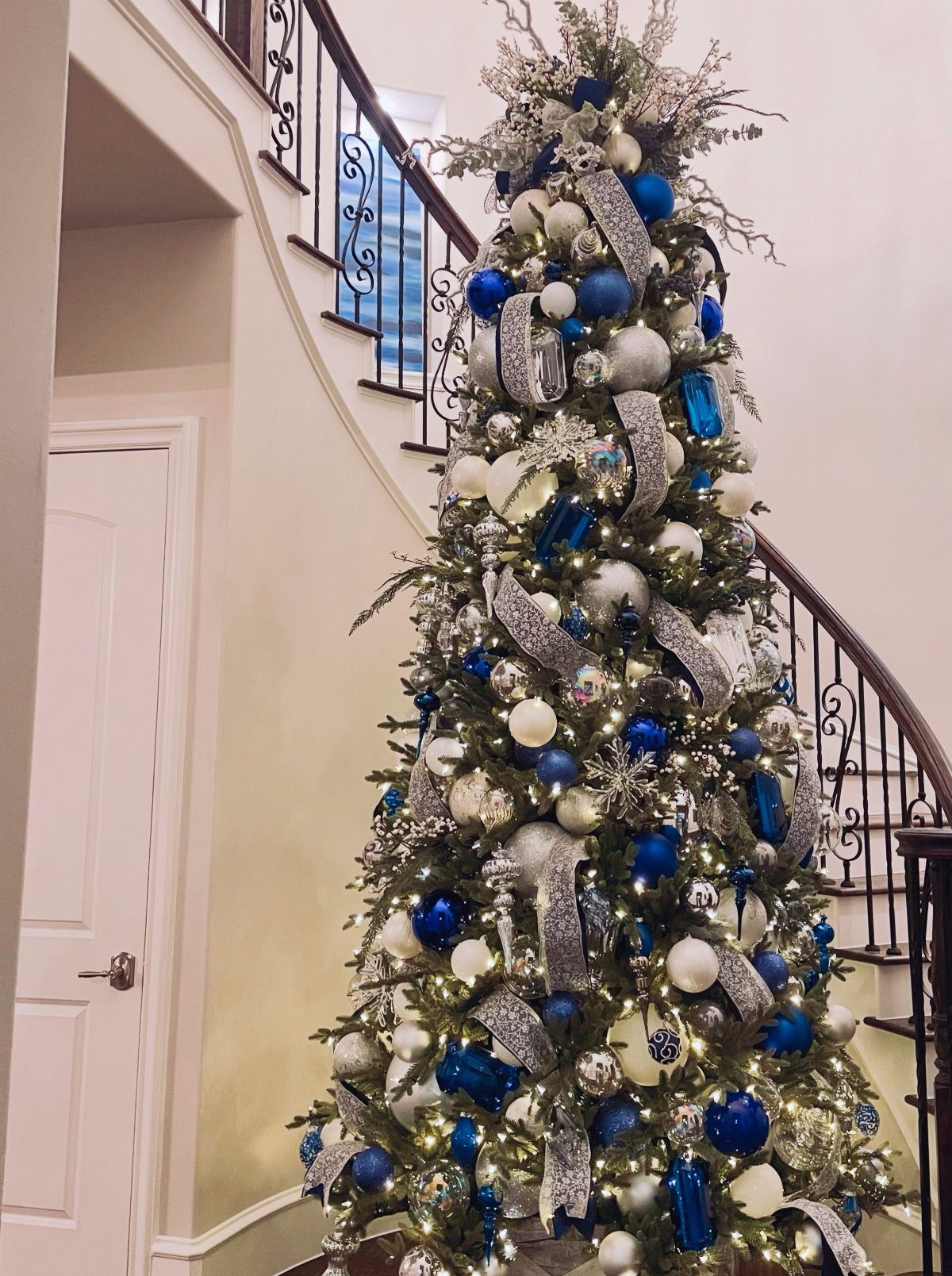 A Christmas tree decorated with blue ornaments and silver ribbon accents next to a staircase.