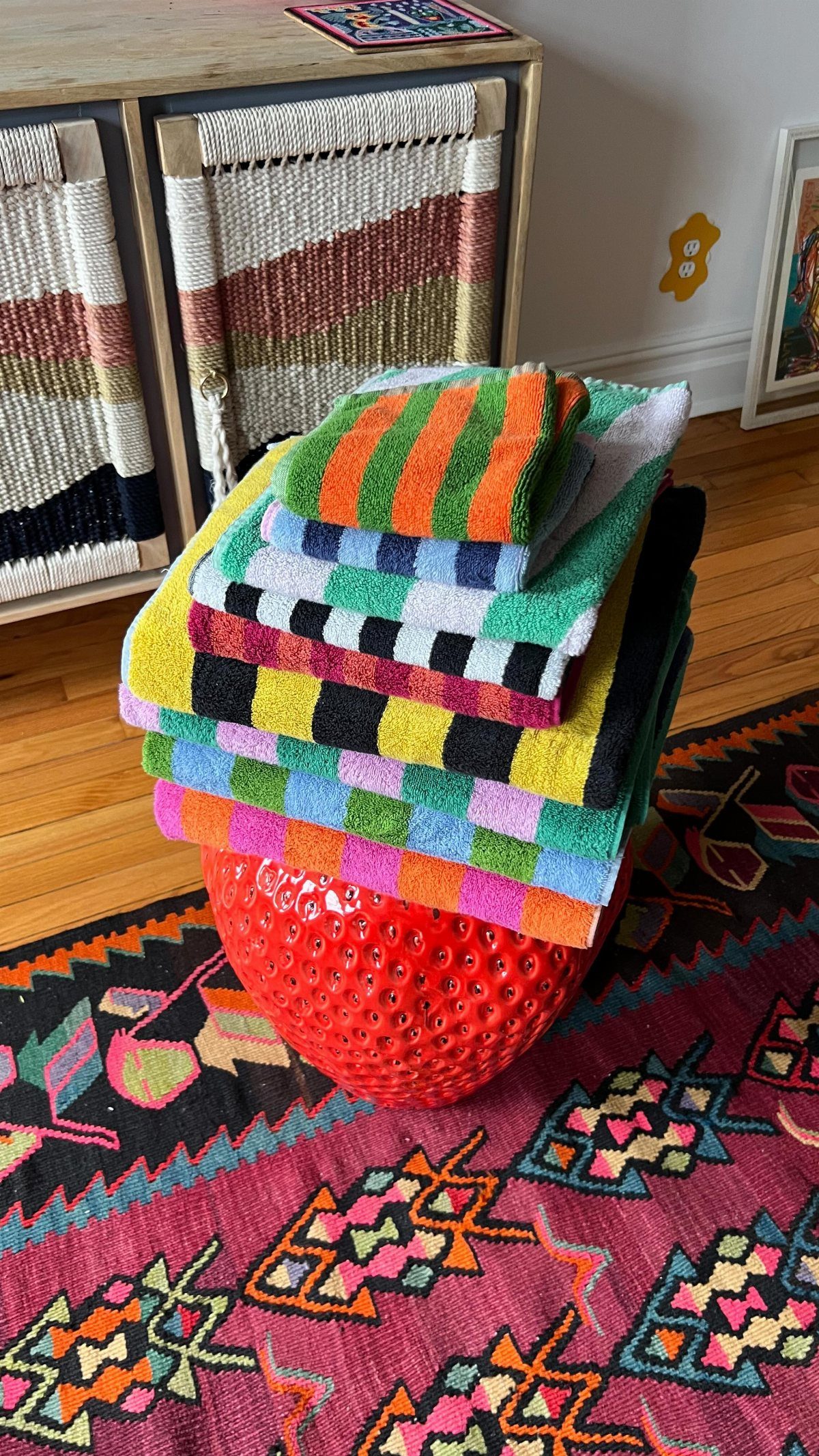 Stacked colorful patterned towels resting on a red ceramic stool over a patterned rug.