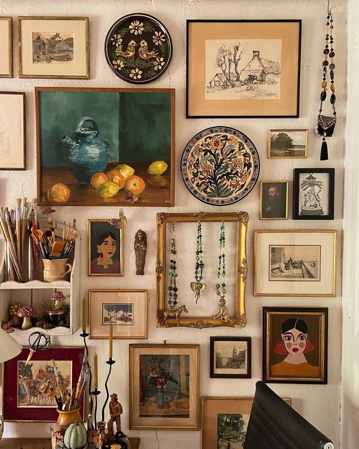 A diverse collection of framed art, decorative plates, and hanging jewelry arranged on a white wall.